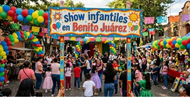 Fotografía con orientación horizontal de una animada fiesta infantil al aire libre en una calle. En primer plano, destaca un gran letrero colorido sostenido por dos postes decorados, con el texto 'show infantiles en Benito Juárez' rodeado de dibujos festivos como soles y globos. Detrás del letrero, hay una gran multitud de niños y adultos disfrutando del evento. La escena está enmarcada por arcos de globos multicolores y tiras de papel picado tradicional que cruzan la calle. Al fondo, se alcanza a ver un pequeño escenario con animadores disfrazados y puestos de comida a los costados, bajo una luz de día soleada.